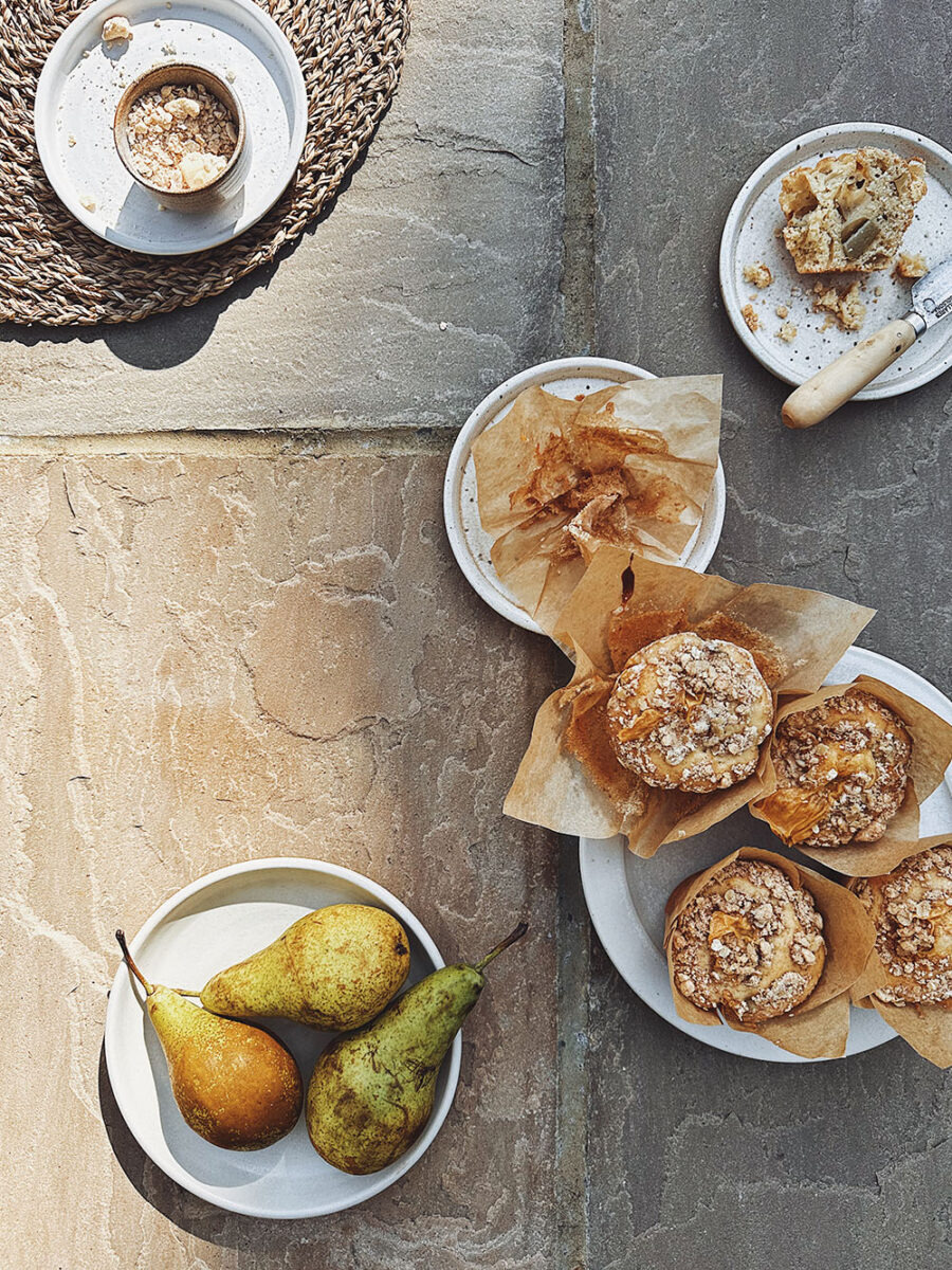 Five small plates are set on a beige stone backdrop, some with pears on and others with crumble and custard muffins on. Scrunched up muffins cases and half eaten muffins can also be seen. 
