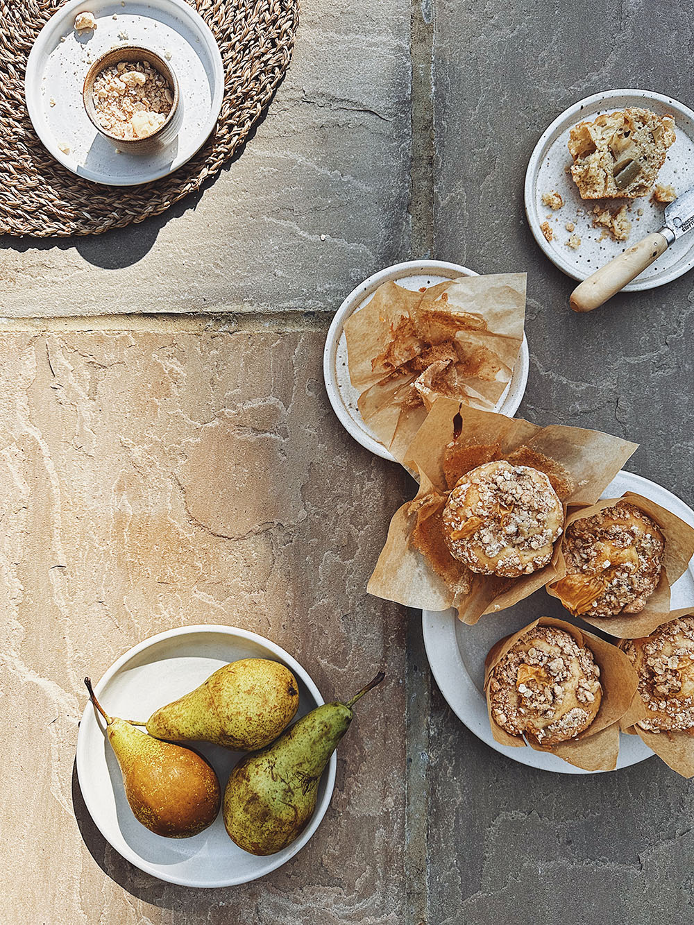 Five small plates are seen on a beige stone backdrop, one is filled with pears and the others with crumble and custard muffins, some half eaten in the sunshine