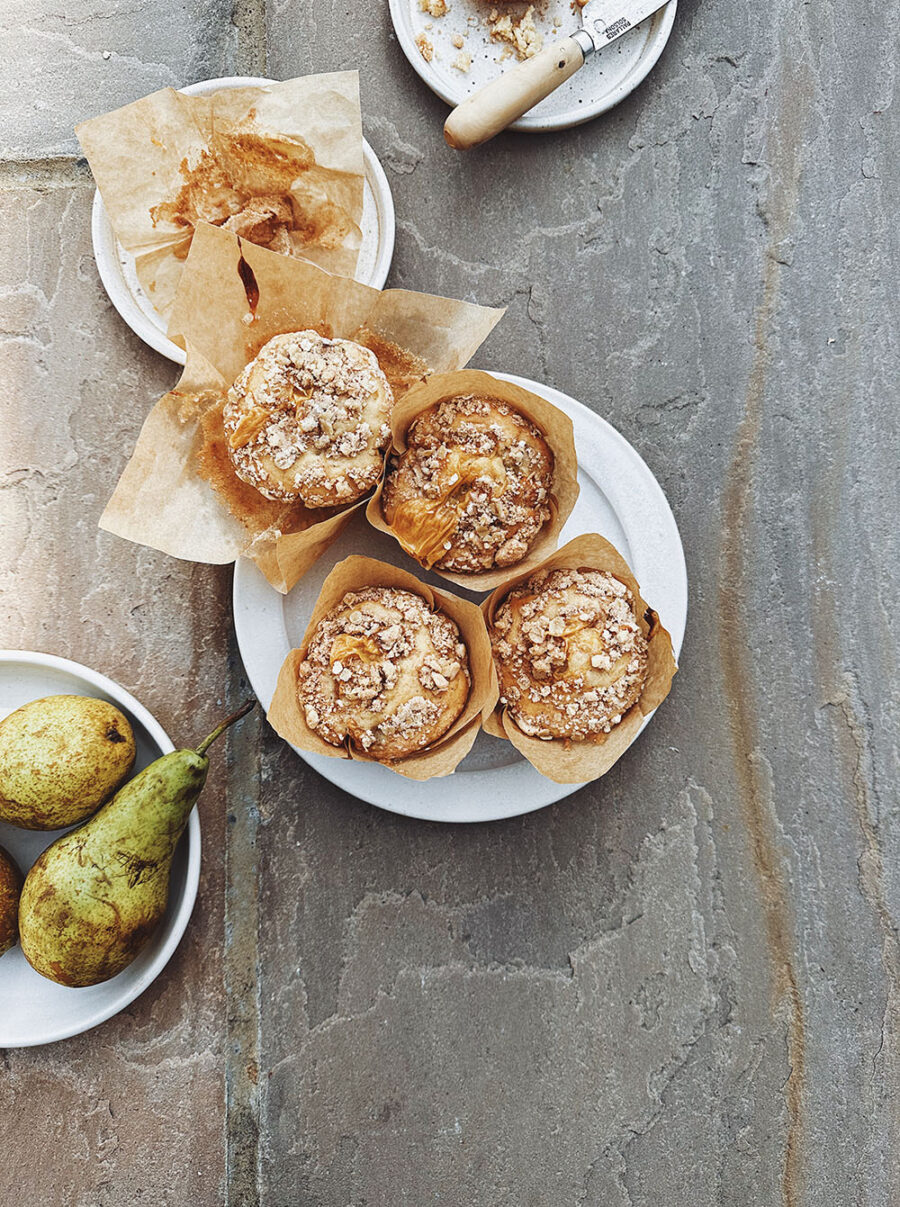 Four crumble and custard muffins on a white plate set on a beige stone backdrop. On the left lower side of the photo, there's a plate of pears and above, speckled plates with muffins cases and crumbs on.