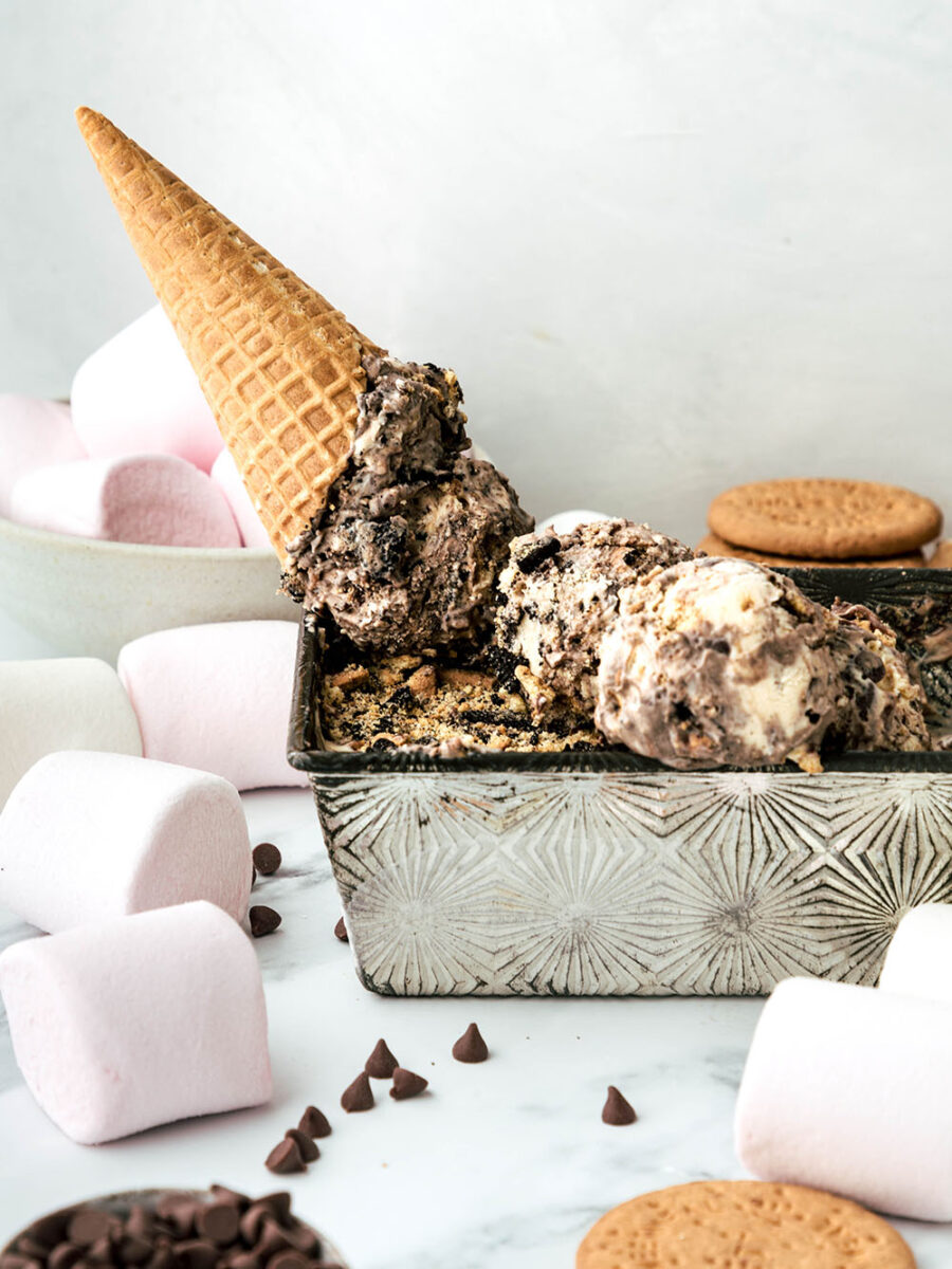 An upturned cone sits in a metal tin of scooped S'mores Ice Cream balls, on top of a marble worktop. Fluffy marshmallows and digestive biscuits can be seen in the background.