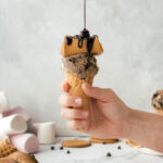 Someone is holding a cone filled with S'mores Ice Cream that's topped with a toasted marshmallow, with chocolate fudge sauce being drizzled on top.