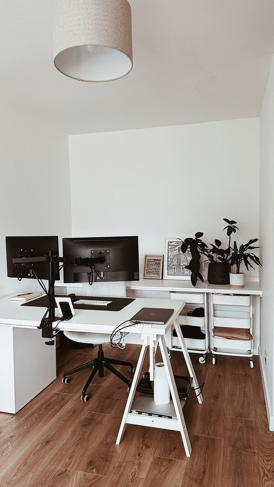 A home studio with wooden floors and white walls. An IKEA desk is positioned against the left wall, with a laptop and two monitors held up by metal arms. There are two trolleys underneath a backboard and some plants in the right hand corner.