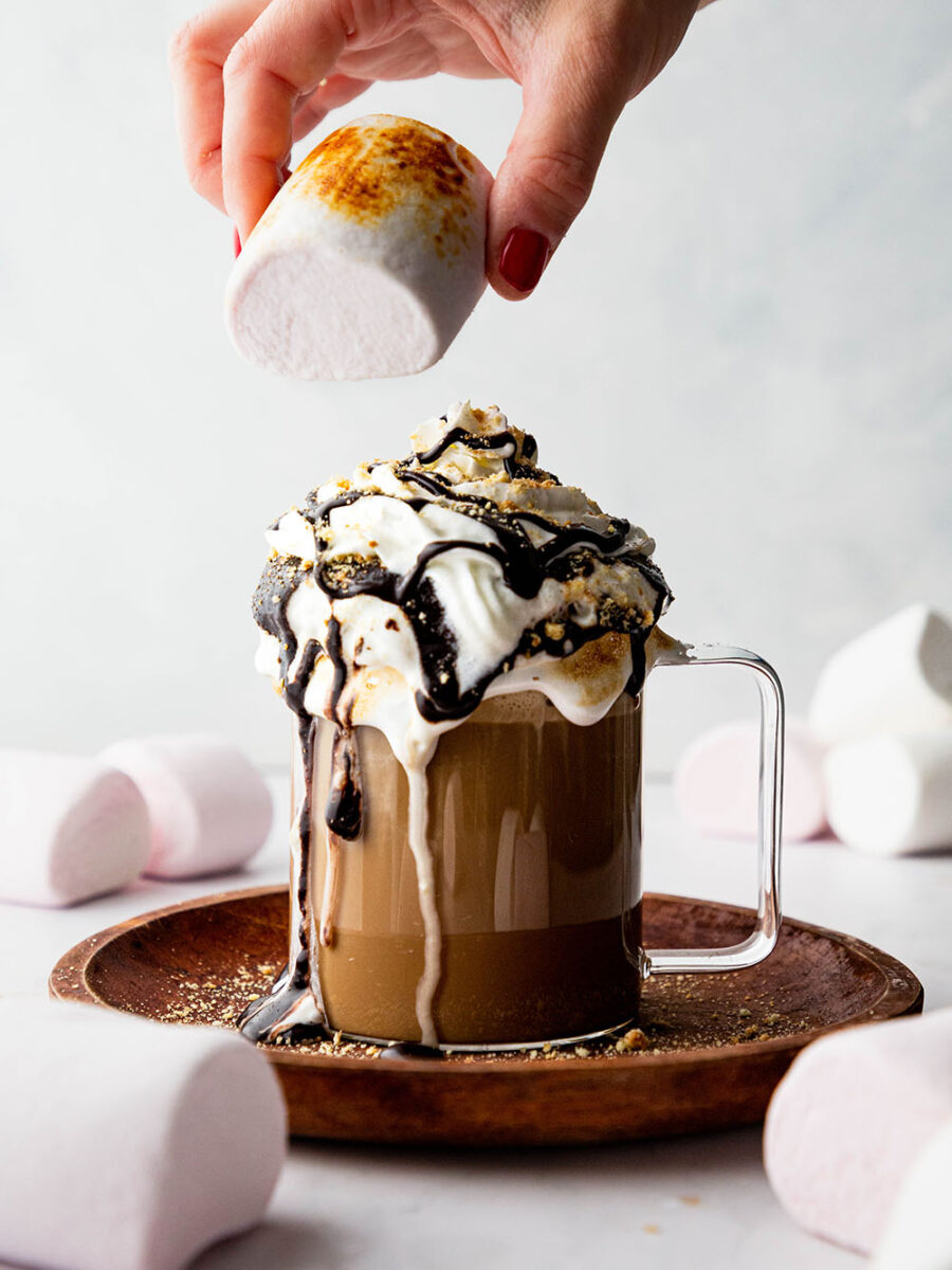 A glass mug of chocolate coffee and marshmallow milk is topped with whipped cream, chocolate sauce and crushed digestive biscuits. The cream is drizzling down the sides of the mug and a hand is about to top the drink with a toasted marshmallow.