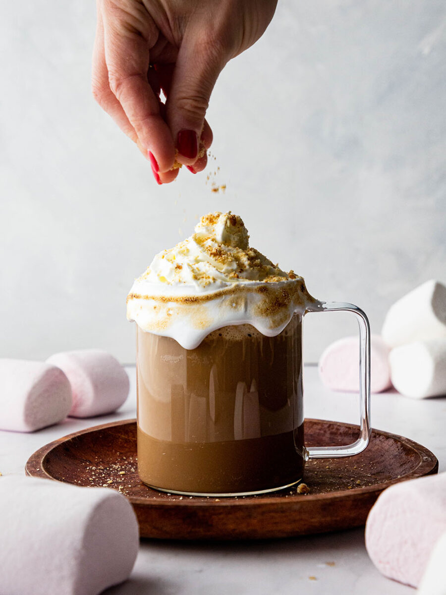 A glass mug of chocolate coffee and marshmallow milk is topped with whipped cream. Marshmallows are scattered on a grey marble backdrop and a hand is sprinkling crushed digestive biscuits over the top of the drink.
