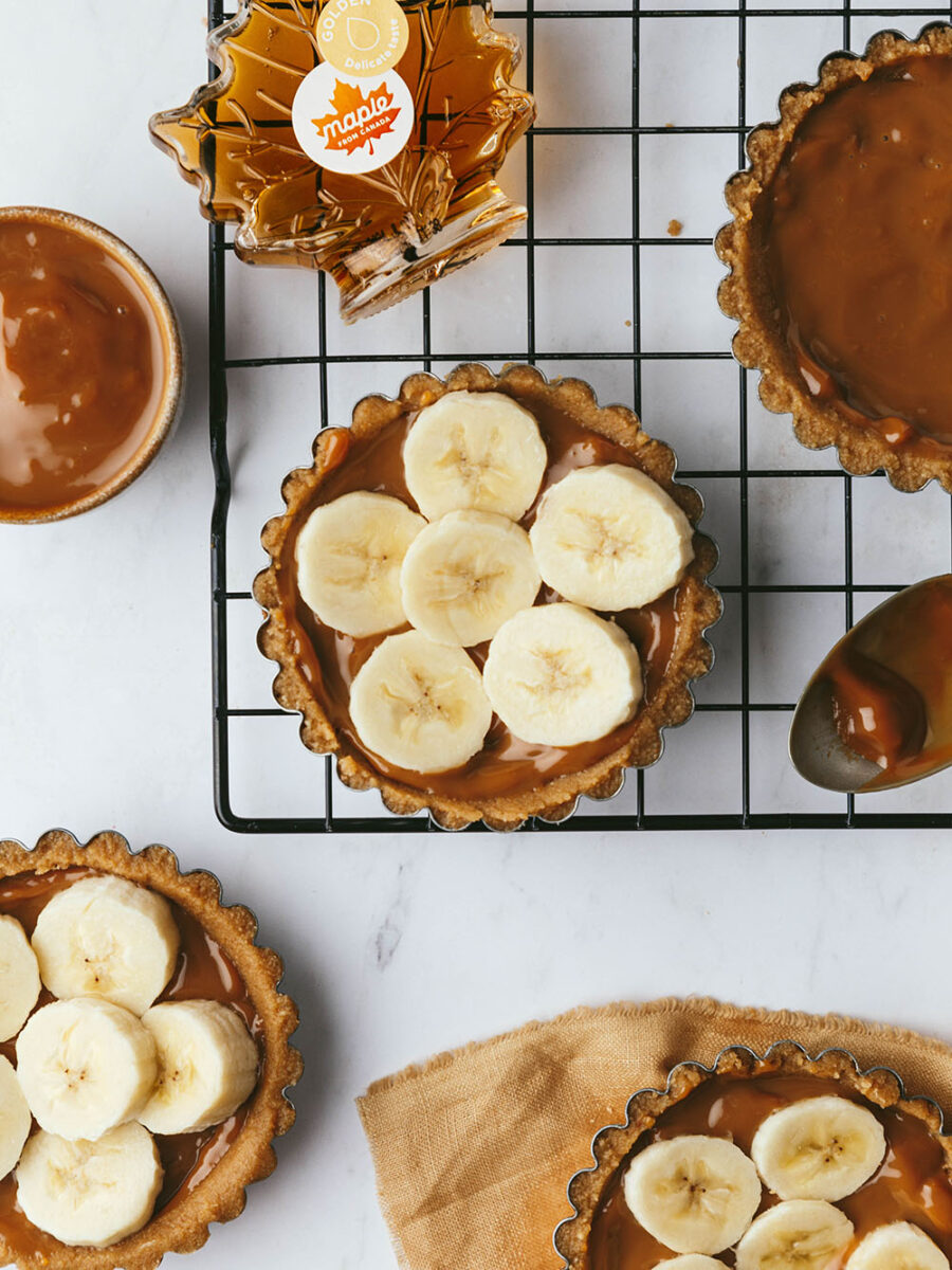Round mini maple Banoffee pies on a marble backdrop with caramel and banana filled biscuit bases. A bottle of maple syrup and a pot of caramel can also be seen, and a sliver of yellow linen is peaking in on the bottom right hand corner.