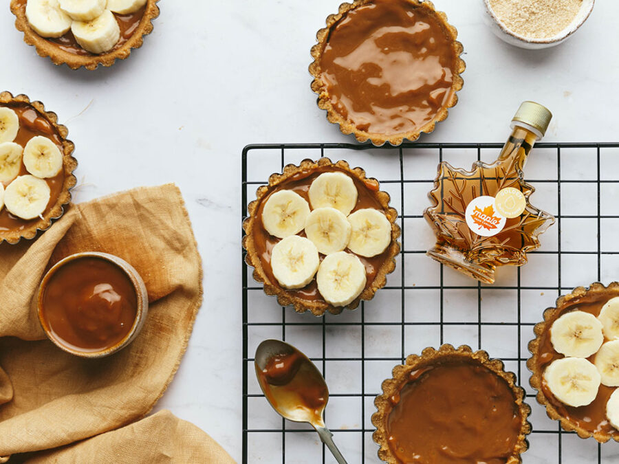 Round mini maple Banoffee pies on a marble backdrop with caramel and banana filled biscuit bases. A bottle of maple syrup and a pot of caramel can also be seen, and a sliver of yellow linen is peaking in on the bottom left hand corner.