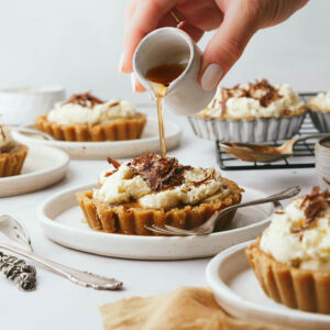 A hand is pouring light maple syrup onto mini maple Banoffee pies which have been placed on neutral coloured ceramic plates and a marble backdrop. A yellow linen peeks into the bottom right hand corner.
