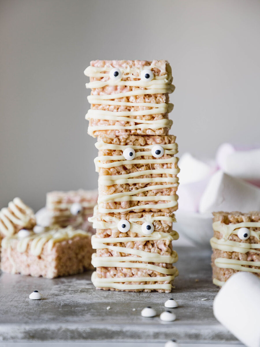 A stack of Rice Krispie marshmallow bars are set on a silver baking tray with more bars and marshmallows in the background. The bars are drizzled in white chocolate and have google eyes on them.