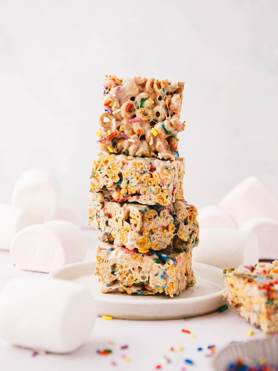 A stack of 4 cereal marshmallow squares can be seen on a neutral backdrop. Marshmallows, funfetti sprinkles and more funfetti marshmallow bars can be seen dotted around.