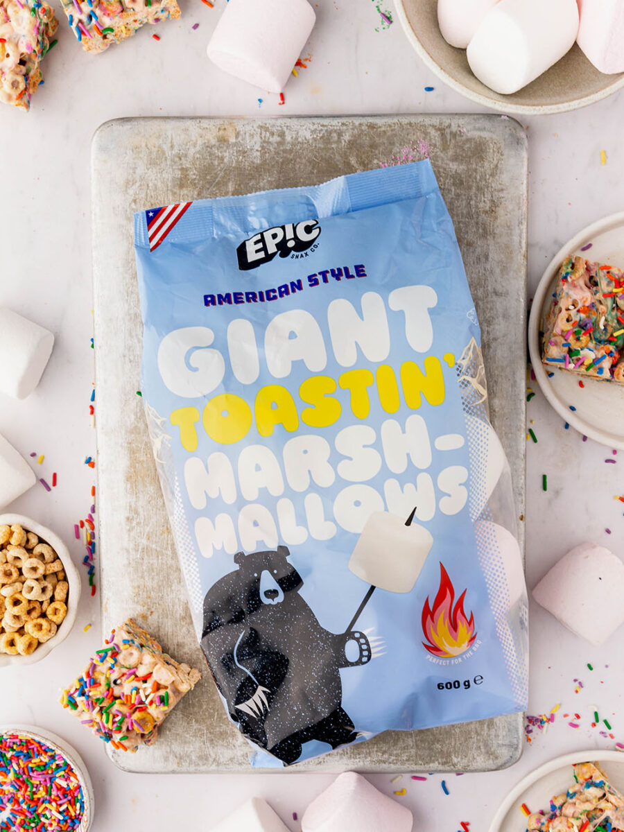 A packet of Epic Snax's Giant Toastin' Marshmallows can be seen on a silver baking tray set on a white neutral backdrop. Funfetti marshmallow bars, funfetti sprinkles, cheerios and marshmallows are dotted around the edges of the frame.