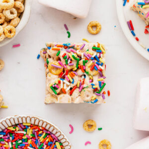 A cereal square sits in the middle of a white backdrop with funfetti sprinkles on top and some in a small bowl in the bottom left hand corner. A bowl of cheerios can be seen in the top left hand corner and marshmallows in the bottom right hand corner.