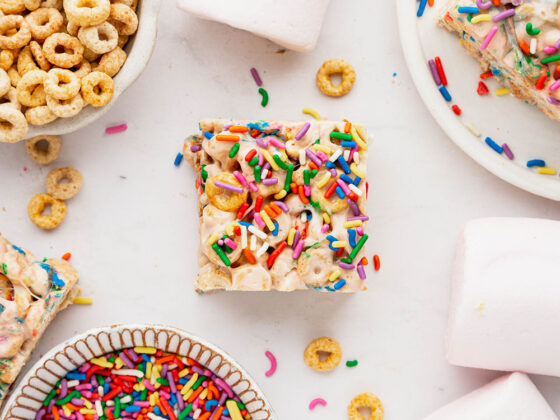 A cereal square sits in the middle of a white backdrop with funfetti sprinkles on top and some in a small bowl in the bottom left hand corner. A bowl of cheerios can be seen in the top left hand corner and marshmallows in the bottom right hand corner.