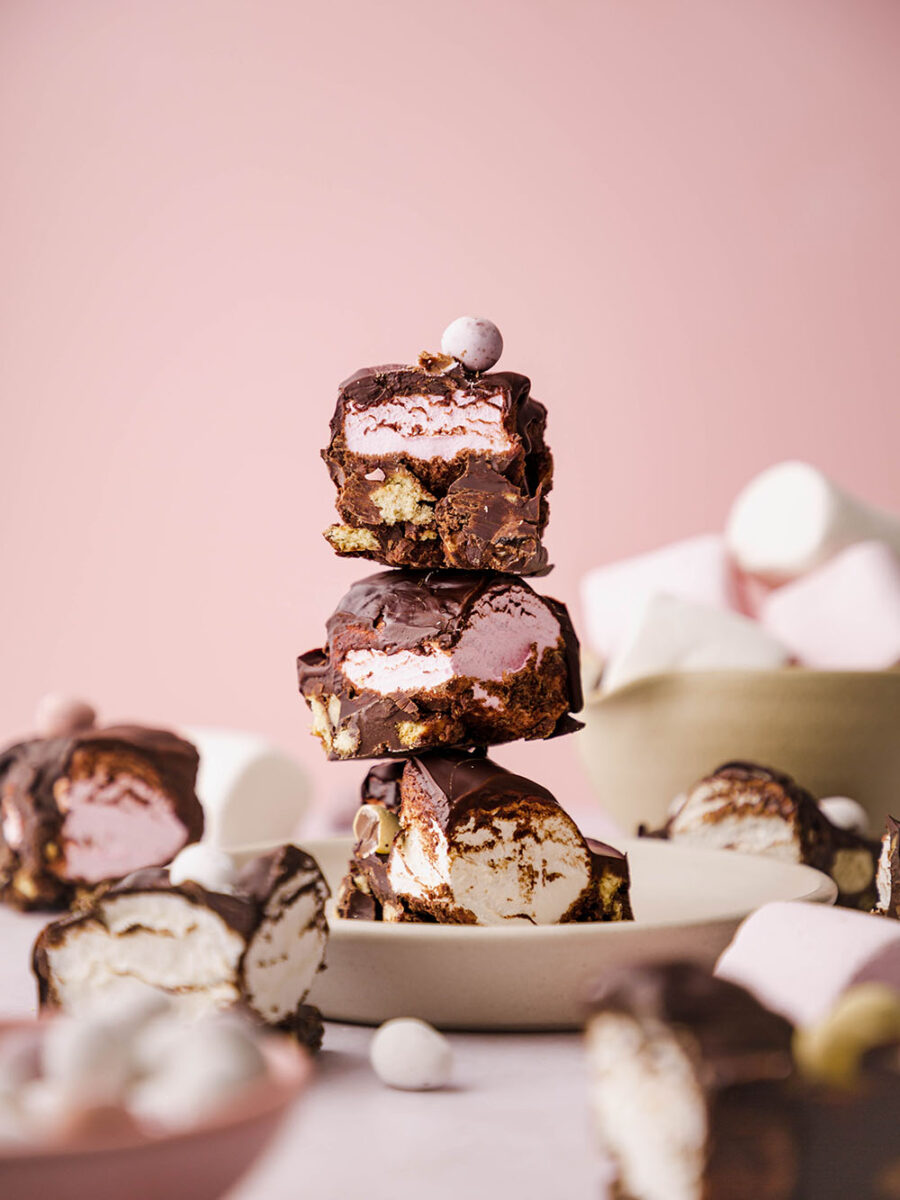 Chocolate covered marshmallow rocky road is stacked up on a plate against a pale pink background. Mini eggs are on top of the squares and more can be seen scattered in the background with marshmallows.