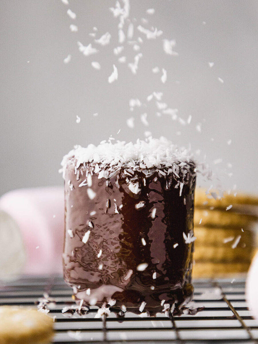 A chocolate coated marshmallow being sprinkled with shredded coconut on a black wire rack. Biscuits and marshmallows can be seen in the background.