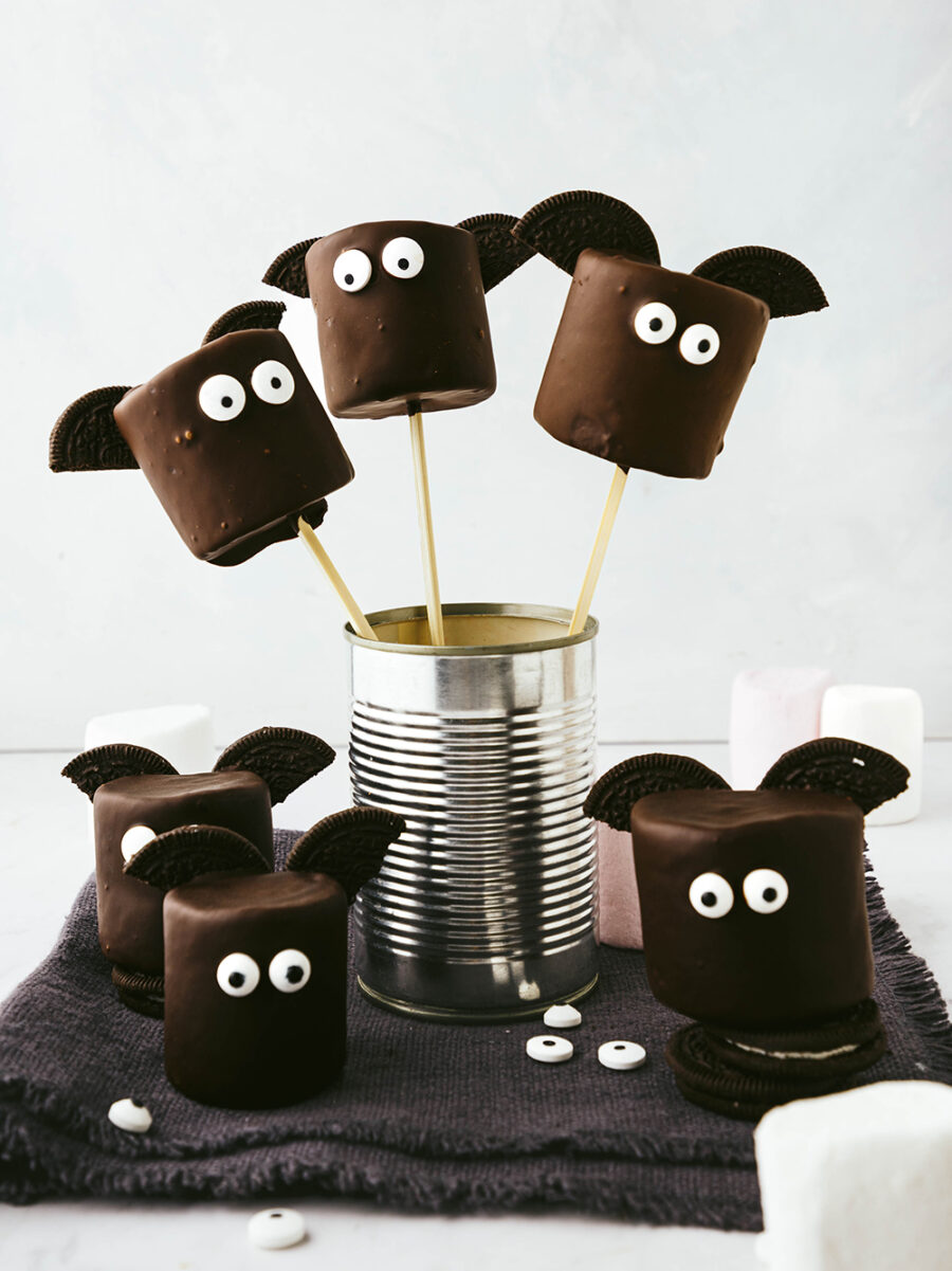 Three chocolate covered marshmallow halloween bats with Oreo halves for wings and edible eyes sit on skewers in an empty metal can. A few more can be seen scattered around on a dark grey fringed napkin against a light grey backdrop.