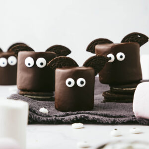 Chocolate covered marshmallows with Oreo halves for wings and edible eyes sit on a dark grey fringed napkin against a light grey backdrop.