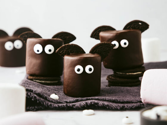 Chocolate covered marshmallow bats with Oreo halves for wings and edible eyes sit on a dark grey fringed napkin against a light grey backdrop.