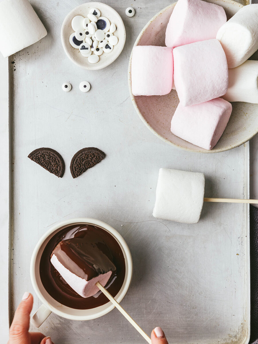 Marshmallow halloween bats: A bowl of marshmallows and a small pinch pot of edible eyes sit on a silver vintage baking tray. Two half of an Oreo are placed on the tray to look like wings and someone is dipping a marshmallow on a skewer into a bowl of dark chocolate with their hands.
