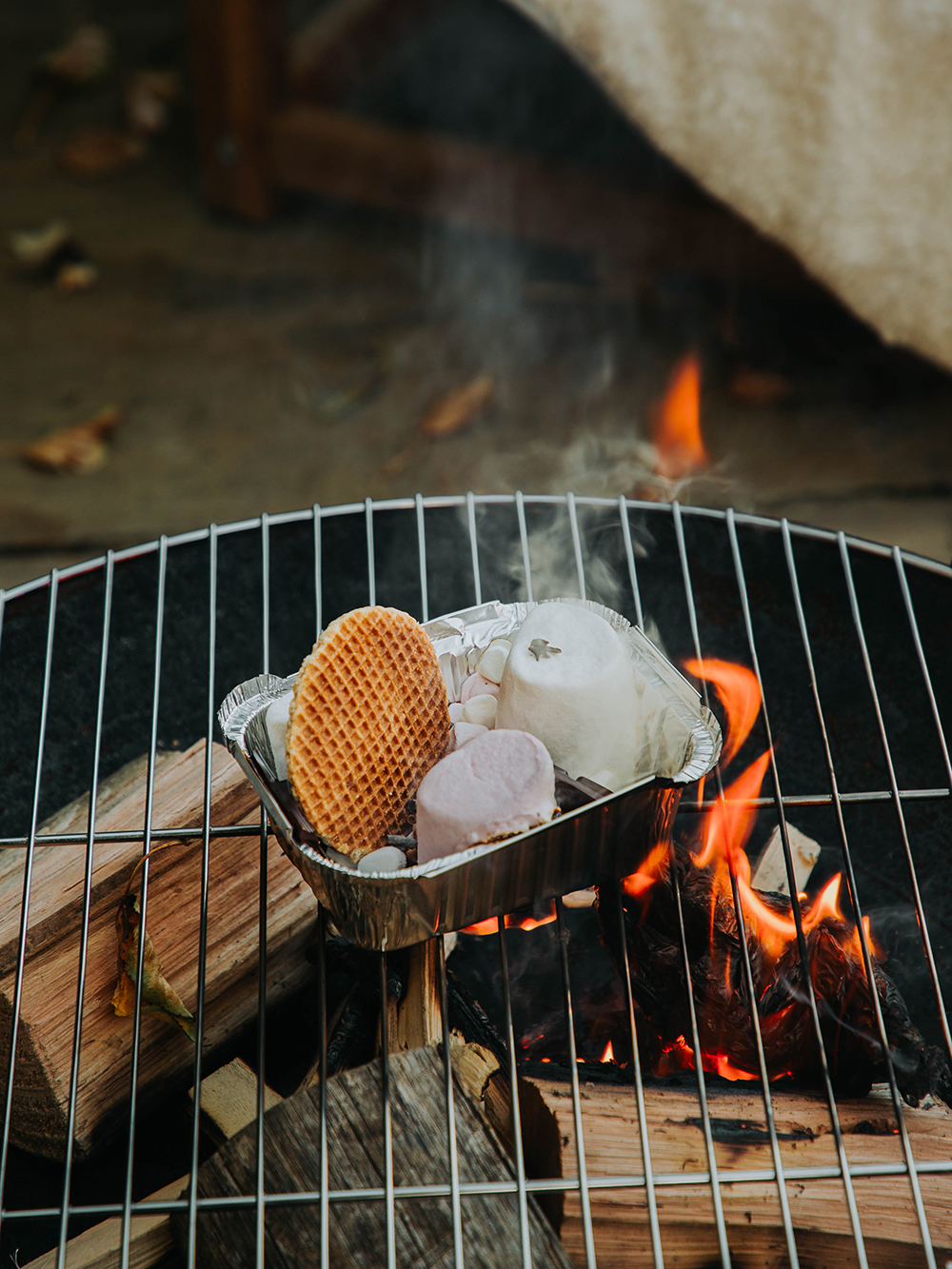 A tin foil box filled with marshmallows, stroopwafels, chocolate and biscuits sits on a wire grate on top of a fire pit.