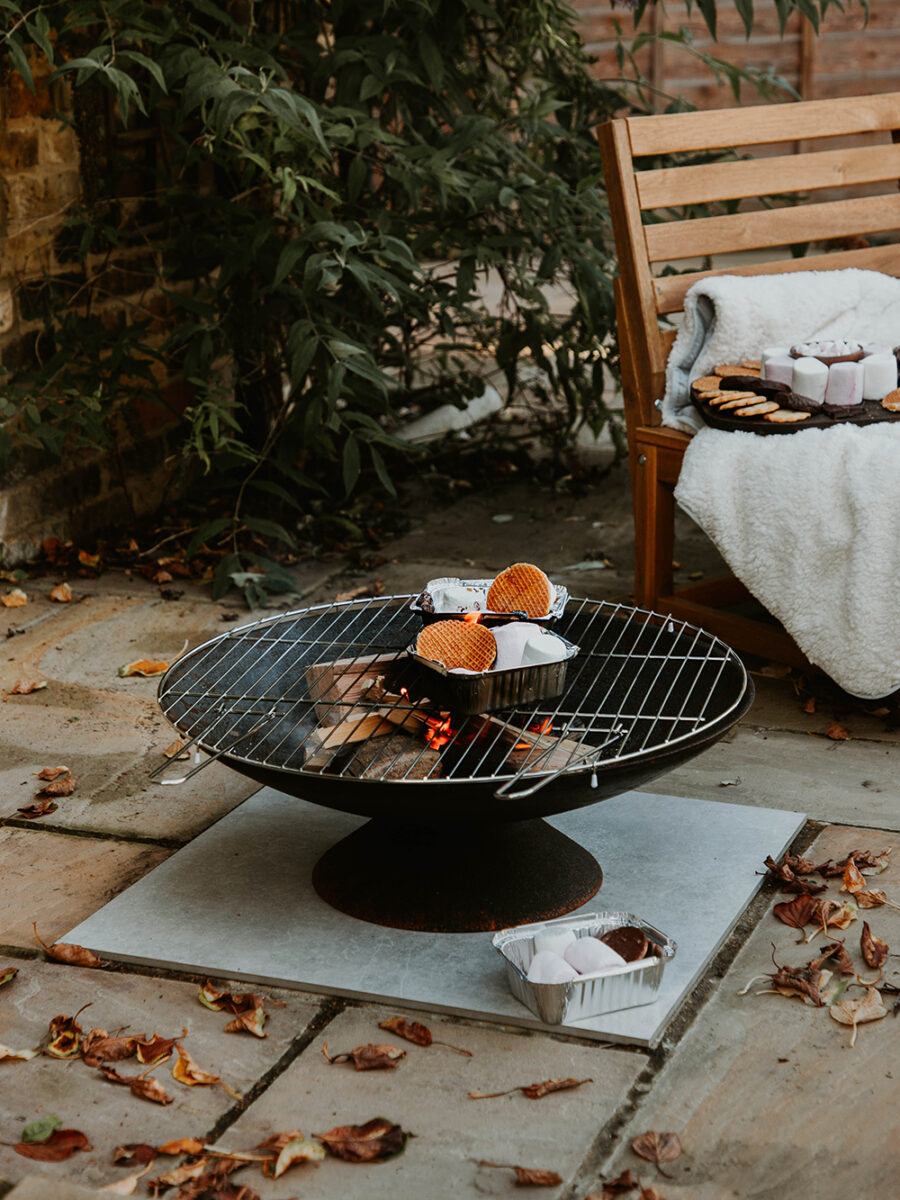 A fire pit on a patio, with flames and smoke. On top there are tin foil boxes filled with marshmallows, stroopwafels, chocolate and biscuits. 