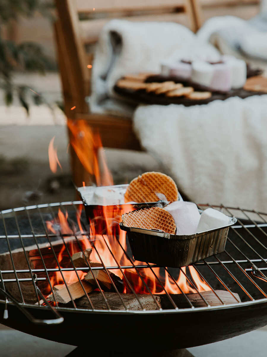 A fire pit with flames and two tin foil boxes on top, filled with marshmallows, stroopwafels, chocolate and biscuits. A bench with a platter is in the background.