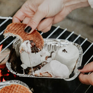 Someone is holding a bitten stroopwafel and is scooping up some melted chocolate and marshmallows from a tin foil box.