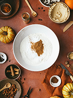 A white mixing bowl with flour and pumpkin spice mix on a terracotta orange stone backdrop. Bowls with icing, and pinch pots with spices lay scattered around and small pumpkins are creeping into the frame.
