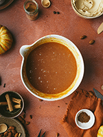 A white mixing bowl with flour and pumpkin spice mix on a terracotta orange stone backdrop. Bowls with icing, and pinch pots with spices lay scattered around and small pumpkins are creeping into the frame.