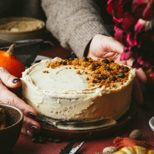 A pumpkin spice latte round cake sits on a wooden plate, with crushed biscuit and coffee beans on top. Two hands are around the cake as if to put it on the table. Ingredients and small pumpkins are scattered around it.