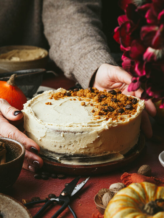 A pumpkin spice latte round cake sits on a wooden plate, with crushed biscuit and coffee beans on top. Two hands are around the cake as if to put it on the table. Ingredients and small pumpkins are scattered around it.