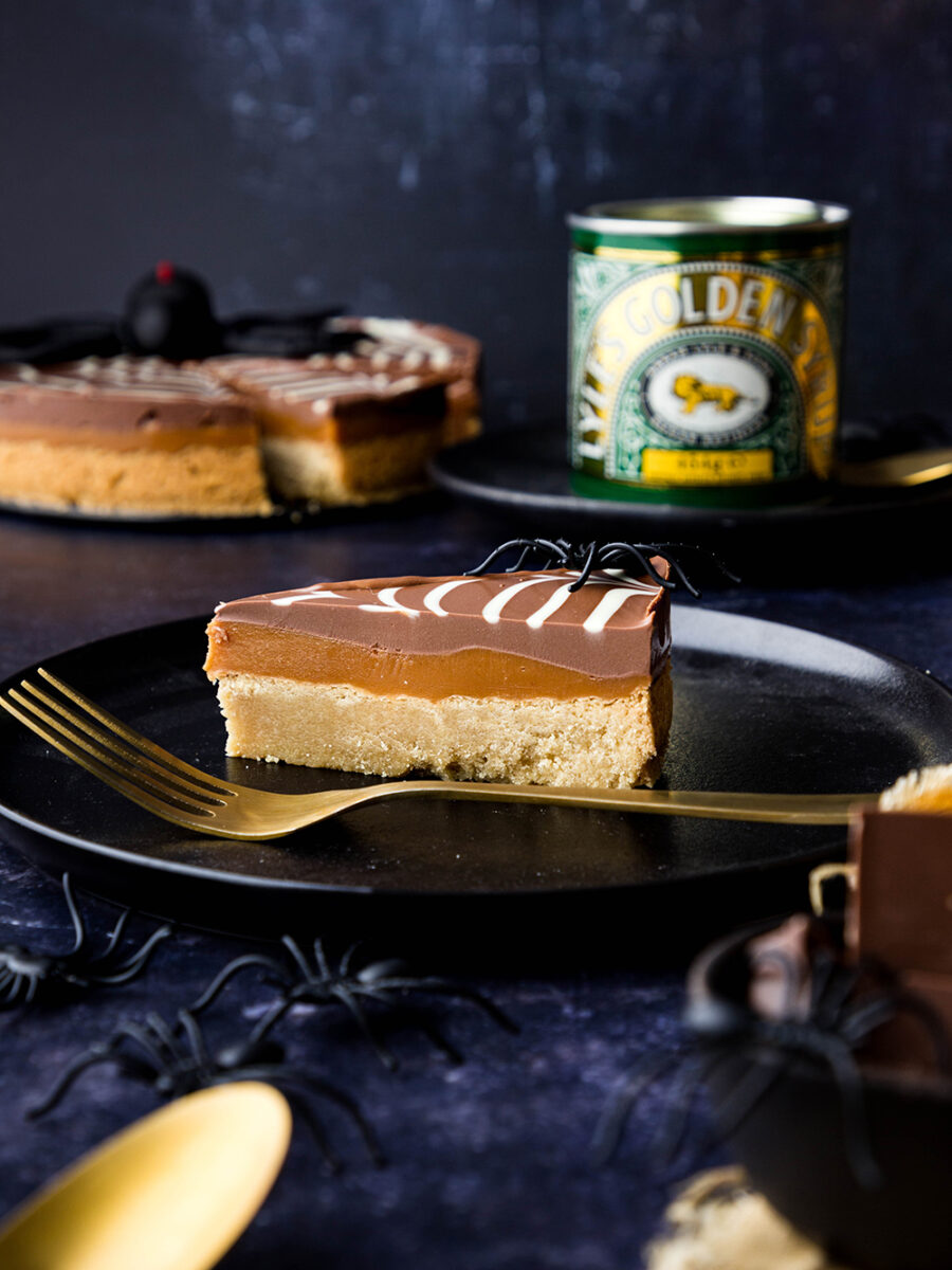 A slice of millionaire's spiderweb shortbread is on a black plate against a dark blue backdrop. You can see the rest of the round shortbread in the background along with a Lyle's golden syrup tin.