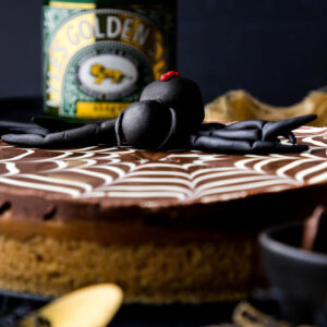 A round millionaire's shortbread with a spiderweb effect chocolate top and a fondant spider is on a dark blue / black background. Little spiders and gold linens and spoons can be seen creeping into the scene. A Lyle's Golden Syrup tin is in the background.