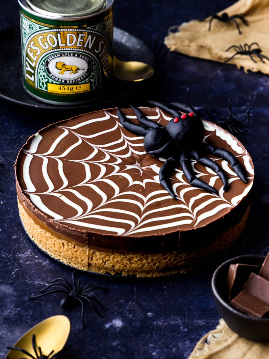 A round millionaire's spiderweb shortbread is set on a dark blue backdrop with a Lyle's golden syrup tin in the background and gold linen, spoons and little black spiders around it. A larger fondant spider sits on top of the dessert.