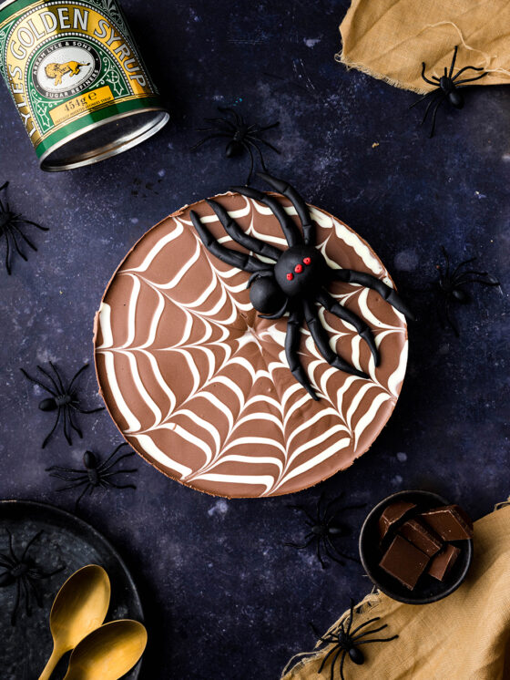 A round millionaire's spiderweb shortbread is sat on a dark blue backdrop. The chocolate topping has been marbled with white chocolate to look like a spiders web and there is a black fondant spider on top. A Lyle's golden syrup tin is in the top left hand corner and there is gold linen, spoons and small black spiders dotted around the dessert.