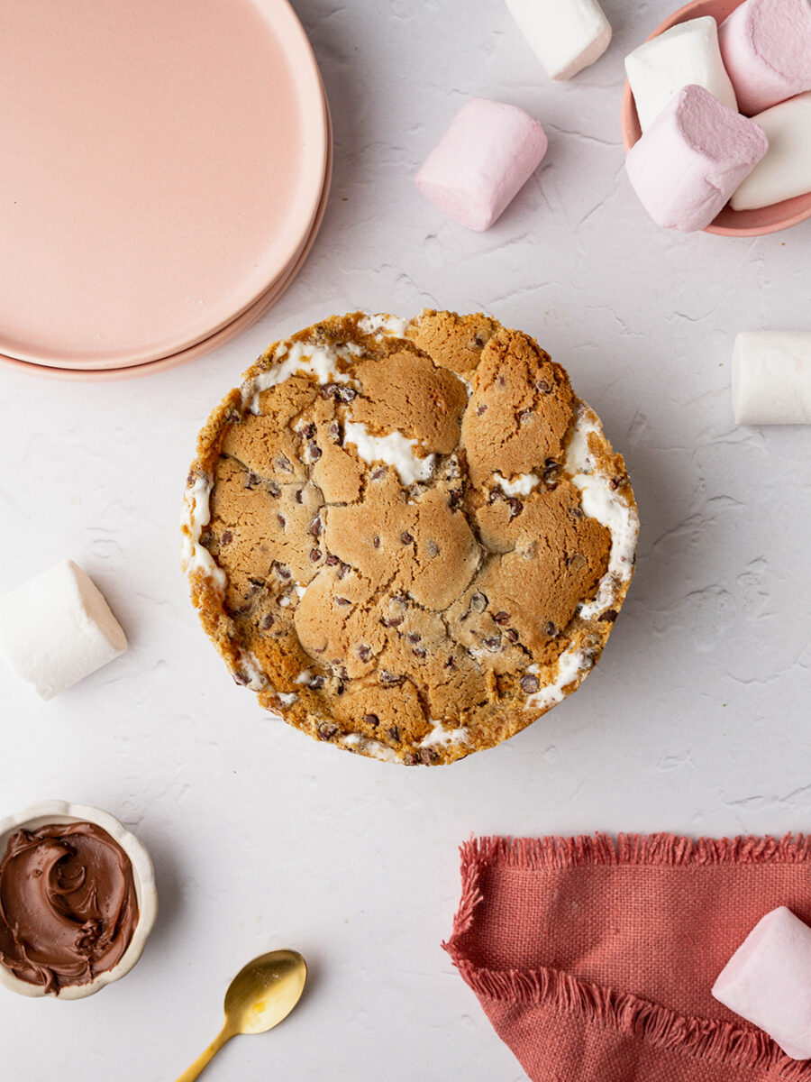 A topdown shot of a cookie pie with marshmallow bursting through the top of the crust, set on a grey marble backdrop. Pink plates can be seen in the top left hand corner, along with a bowl of marshmallows scattered in the top right hand corner. A peachy pink linen frayed napkin peeks into the frame on the bottom right hand side, along with a gold spoon and a small bowl of chocolate spread on the bottom left.