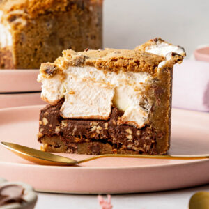 A slice of Marshmallow Cookie Pie filled with marshmallow and chocolate hazelnut spread filling on a pink plate with a gold spoon next to it. The rest of the cookie pie can be seen in the background along with marshmallow in a bowl and scattered on a grey marble backdrop. A peachy pink frayed linen napkin peeks into the bottom right hand corner, along with a golden spoon and a small bowl of chocolate hazelnut spread in the bottom left.