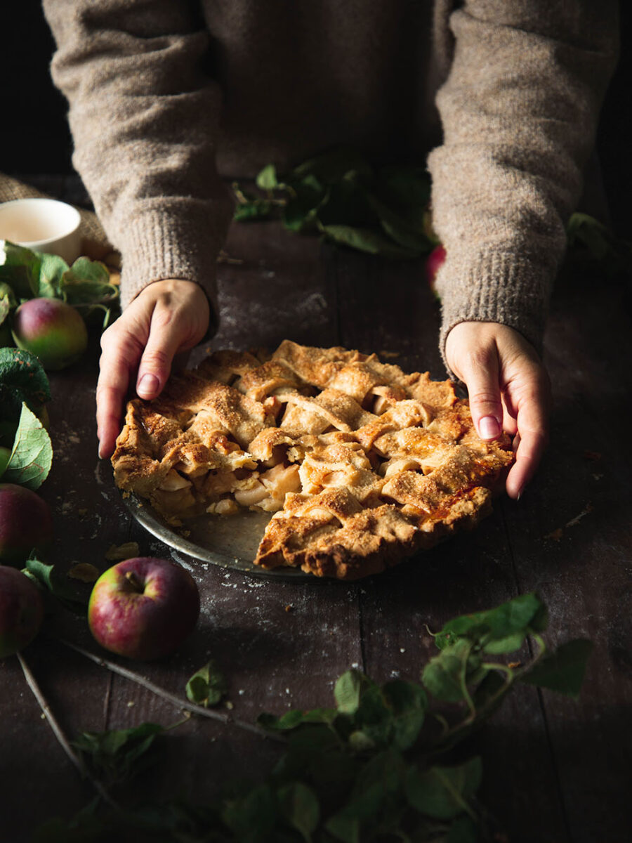 A dark and moody scene with a pallet wood table and a latticed golden apple pie set upon it. Apples and leaves are scatted around it. A person wearing a light brown jump is gently placing the pie down.