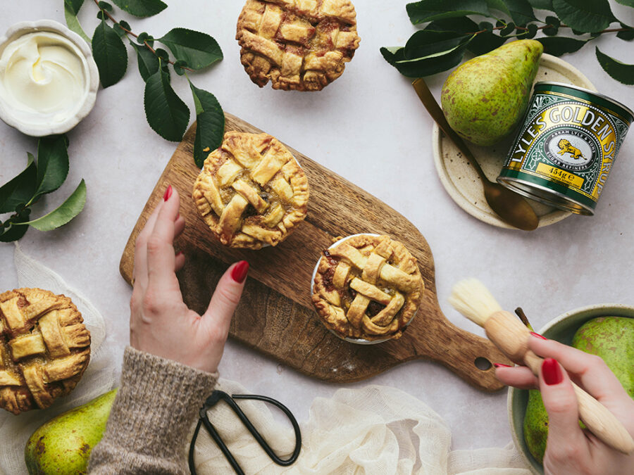A topdown shot of pear, pecan and syrup pies on a wooden board. There's a tin of Lyle's Golden Syrup, a bowl of crème fraîche, pears and leaves scattered around. The backdrop is a marble grey. Some hands are reaching into the shot to glaze the pies with a pastry brush and milk. 