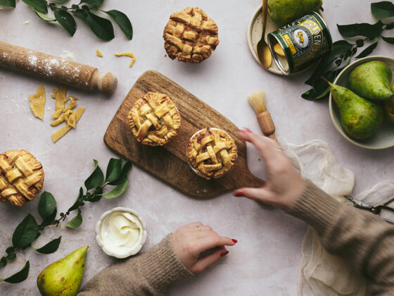 A topdown shot of pear, pecan and syrup pies on a wooden board. There's a tin of Lyle's Golden Syrup, a bowl of crème fraîche, pastry cuttings and a rolling pin, pears and leaves scattered around. The backdrop is a marble grey. Hands are reaching into the shot to take one of the pies off the table.