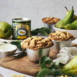 A pear, pecan and syrup pie on a wooden board. There's a tin of Lyle's Golden Syrup in the background, pastry cuttings, pears and leaves scattered around. The backdrop is a marble grey.