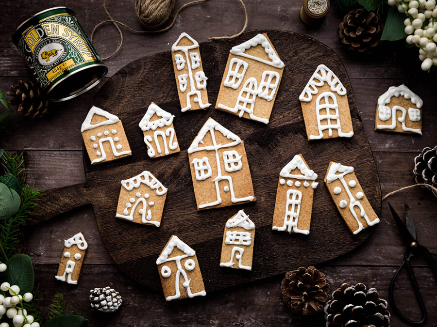 Iced gingerbread houses on a wooden board. Festive christmas foliage surrounds the scene and a Lyle's Golden Syrup tin can be seen in the top left hand corner.