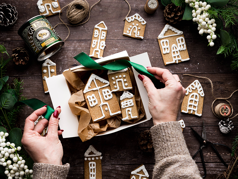 A white box filled with iced gingerbread houses on a wooden table. Festive christmas foliage surrounds the scene and more gingerbread houses are scattered on the table. Someone is reaching in with a green satin ribbon to tie up the box.