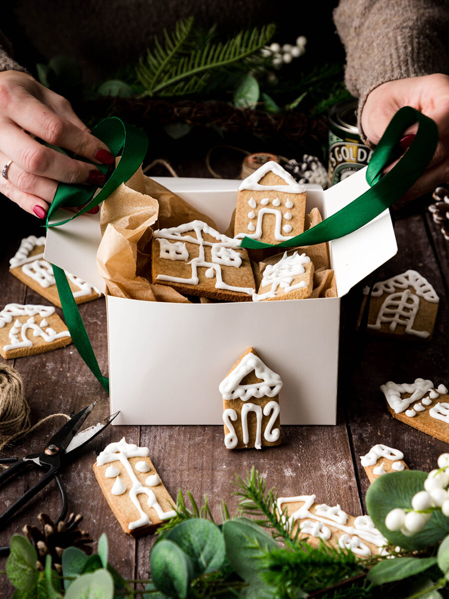 A white box filled with iced gingerbread houses on a wooden table. Festive christmas foliage surrounds the scene and more gingerbread houses are scattered on the table. Someone is reaching in with a green satin ribbon to tie up the box.
