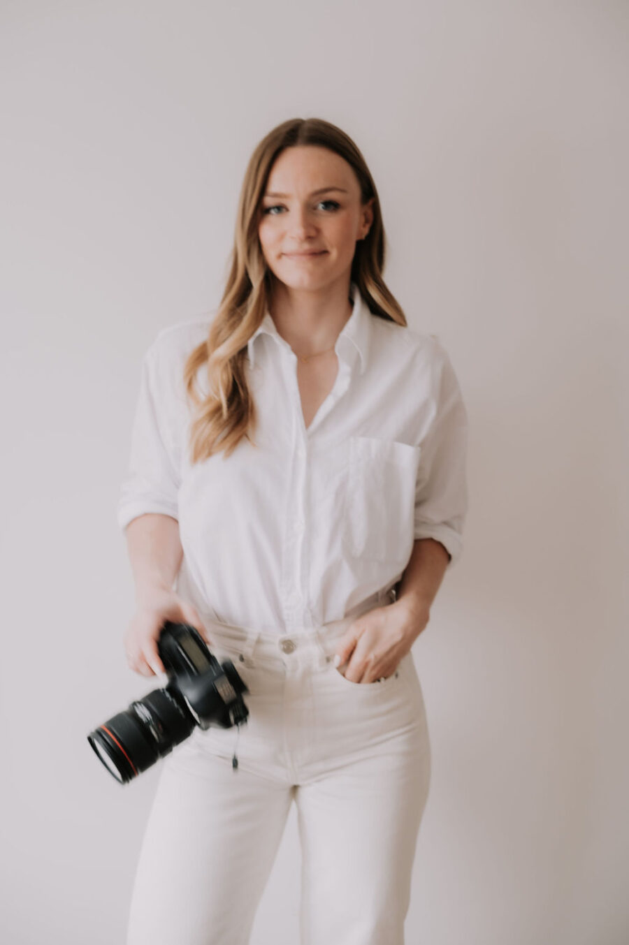 Part-Time Assistant Role: blonde girl standing against white backdrop with white shirt and cream jeans, hand in pocket, holding a camera.