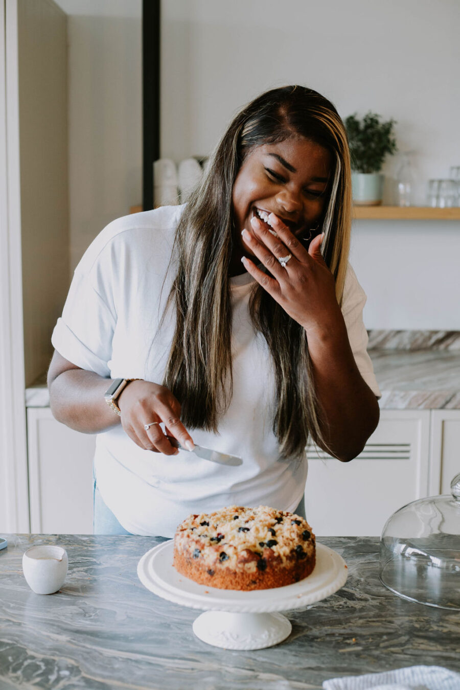 Remi laughing as she cuts into a blueberry cake on white cake stand. Shot in a neutral coloured kitchen with marble tops.